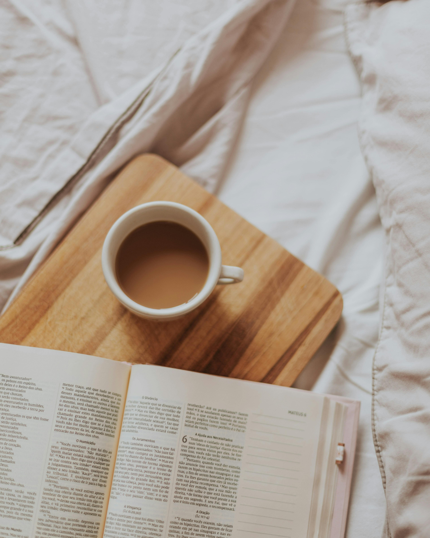 A cup of coffee resting on a wooden board atop rumpled linen sheets, beside an open book in morning light